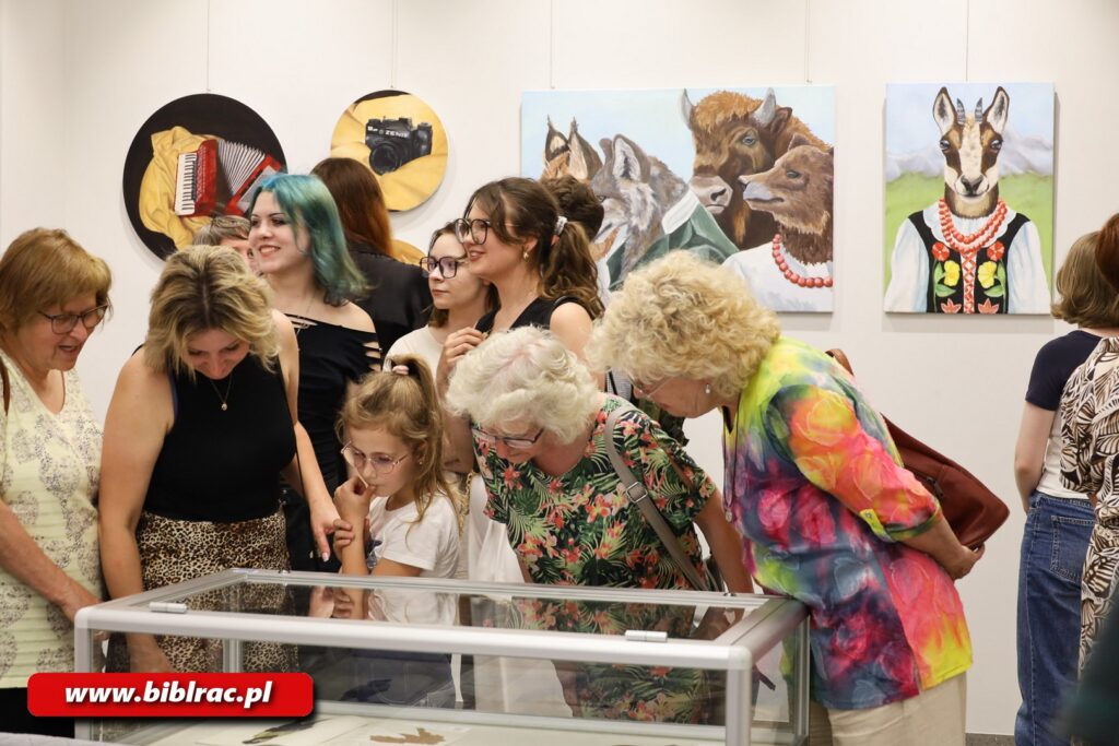 Cztery kobiety i jedna dziewczynka w wieku 7-9 lat pochylające się nad szklaną gablotą wystawienniczą. W gablocie jakieś płaskie eksponaty (grafiki?). Na dalszym planie, za kobietami i dziewczynką przechodzą stoją trzy dziewczyny w wieku nastoletnim. Jeszcze dalej kilka osób zwróconych tyłem do aparatu (a przodem do ściany, na której wiszą obrazy).