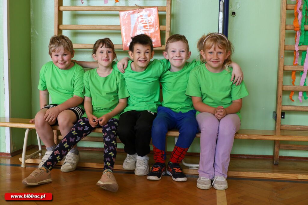 Na zdjęciu grupa pięciorga dzieci, siedzących na długiej ławeczce na sali gimnastyczne, zwróconych twarzami do aparatu, uśmiechających się, obejmujących się rękami. Są to trzej chłopcy i dwie dziewczynki. Wszyscy mają jasnozielone t-shirty bez nadruków i sportowe obuwie. Większość z nich ma założone spodnie dresowe / legginsy, jeden chłopczyk (z lewej) ma krótkie spodenki. Za nimi bladozielona ściana, do ściany zamontowana drewniana drabinka gimnastyczna, do drabinki przymocowany jakiś pomarańczowy niewielki transparent.