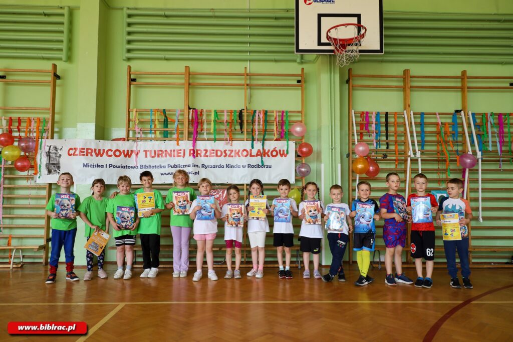 Na zdjęciu grupa 14 dzieci (większość uśmiechnięta) stojąca w rzędzie na sali gimnastycznej. W rękach trzymają prawdopodobnie nagrody wyglądające na czasopisma lub książki z aktywnościami formatu mniej więcej A4. Piątka dzieci w jasnozielonych t-shortach, szóstka w białych t-shirtach, czwórka w strojach wielobarwnych. Wszystcy ubrani na sportowo. Za nimi bladozielona ściana z drabinkami gimnastycznymi i zawieszonym banerem "Czerwcowy turniej przedszkolaków" Na dziećmi na ścianie kosz do koszykówki.