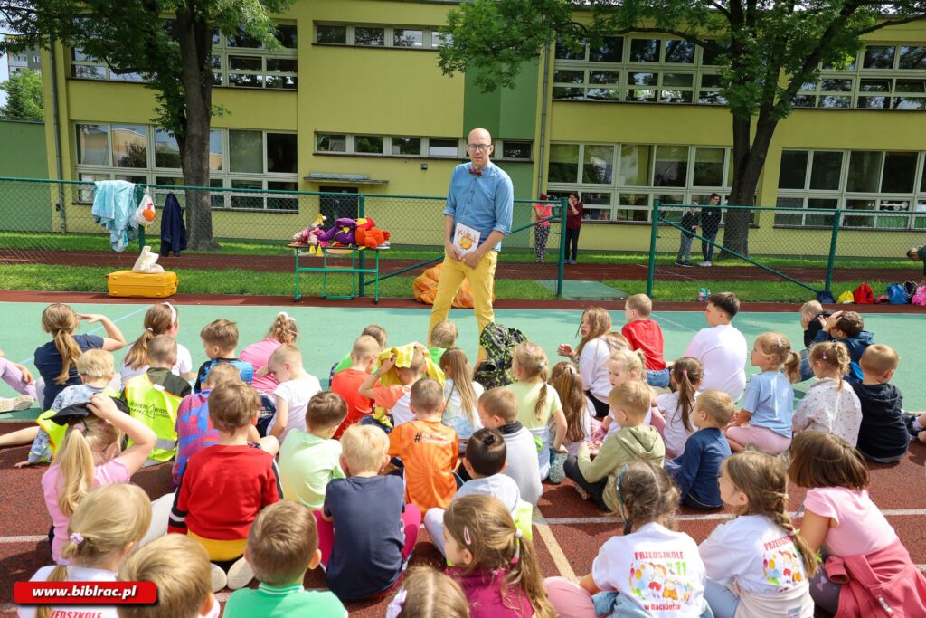 Na zdjęciu na pierwszym planie grupa ok 30 dzieci (prawdopodobnie na boisku szkolnym), siedzących siadem skrzyżnym, zwróconych tyłem do fotografującego,z kolei przodem do mężczyzny stojącego przodem do dzieci (i do aparatu). W tle kosz do koszykówki i fragment budynku (prawdopodobnie szkolnego). Mężczyzna na nosie ma okulary z niebieską ramką, zgolone włosy na głowie, nosi żółte spodnie, niebieską koszulę. Mówi coś do dzieci, w ręce trzyma książkę prawdopodobnie o tytule "Kura". Za mężczyzną ogrodzenie boiska szkolnego, pod i na ogrodzeniu różne rekwizyty (maskotki, stroje). W oddali, za ogrodzeniem, czworo dzieci, za nimi żółto-zielony budynek (prawdopodobnie szkolny).