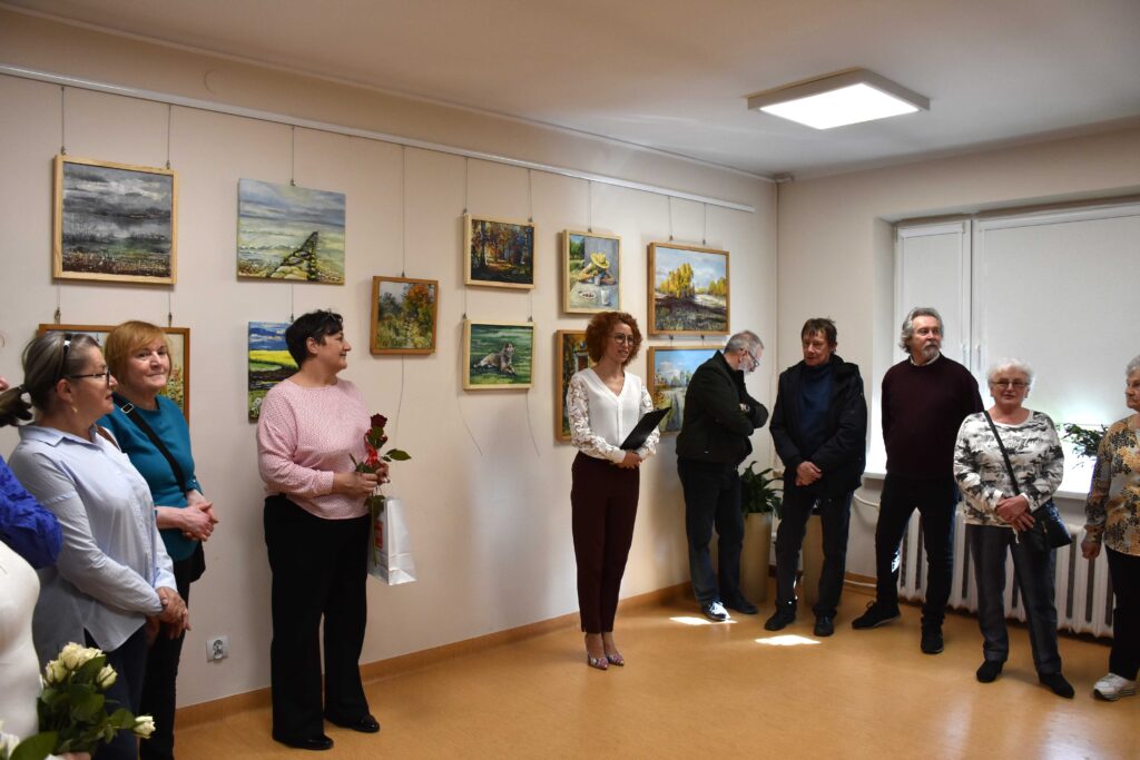 Grupa ludzi stojąca na tle ściany, na której są zawieszone różne obrazy (głównie krajobrazy). Ludzie w wieku średnim i starszym, mężczyźni i kobiety, w sumie 8 osób plus kilka osób niemieszczących się w kadrze. Pośrodku dyrektorka biblioteki (kręcone włosy prawie do ramion, okulary, jasna bluzka, czarne spodnie), przemawiająca i trzymająca podkładkę pod kartkę A4. Obok kobieta w bladoróżowej bluzce/swetrze trzymająca kwiaty i torbę prezentową.