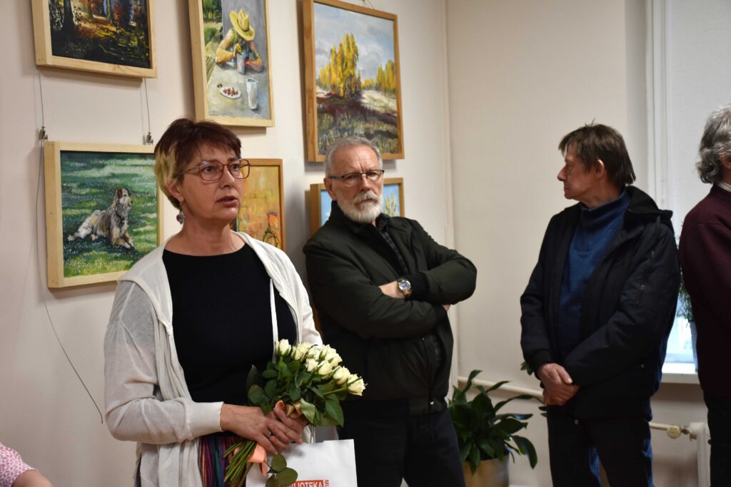 Na zdjęciu na pierwszym planie kobieta (ok 55 lat) o krótkich włosach, w okularach, trzymająca bukiet białych kwiatów ciętych i jakąś torebkę prezentową, na dalszym planie mężczyzna w wieku ok 60 lat z siwą brodą i w okularach, obok niego mężczyzna w podobnym wieku spoglądający na obrazy wiszące na ścianie.