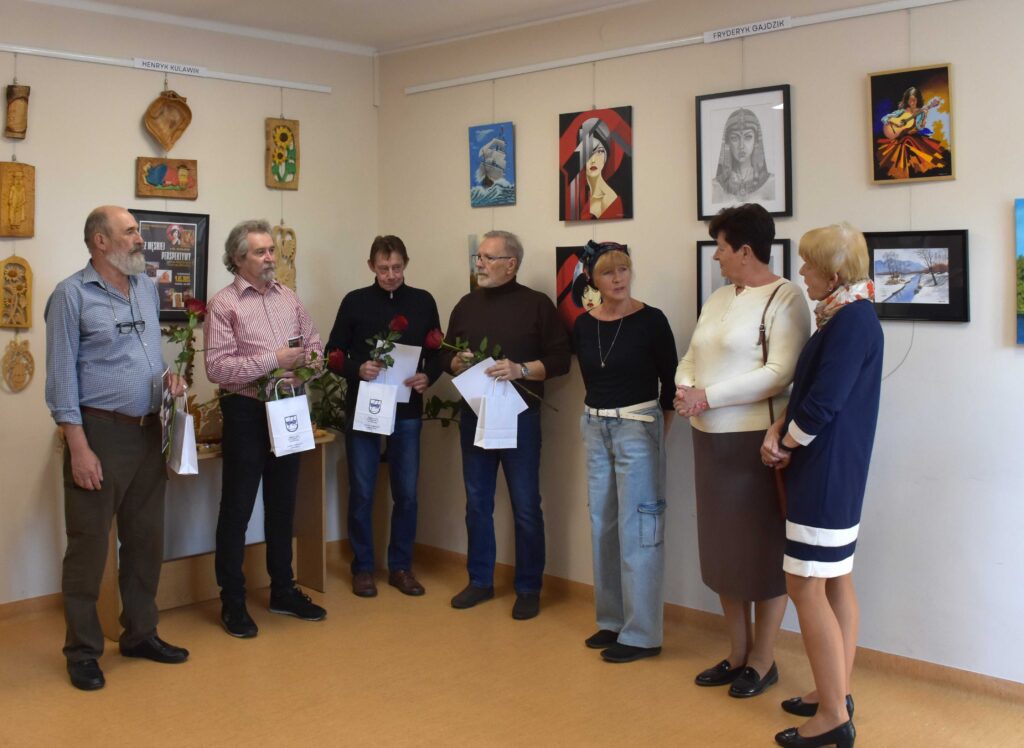 Na zdjęciu czterech mężczyzn i trzy kobiety. Wszyscy ustawieni na tle ściany z obrazami, grafikami i płaskorzeźbami. Mężczyźni trzymają w dłoniach białe torebki prezentowe, róże oraz dyplomy.