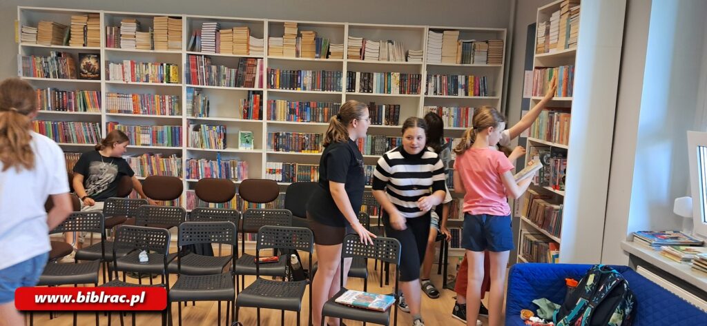 Na zdjęciu dziewczyny w wieku, 12-14 lat, stoją w bibliotece, rozmawiają, ale skupione są na regałach z książkami. Jedna ze stojących dziewczyn sięga do książki na regale, dwie osoby siedzą na krześle, widać, że jedna z nich tez spogląda na regał. Dziewczyny ubrane letnio (krótkie rękawki).