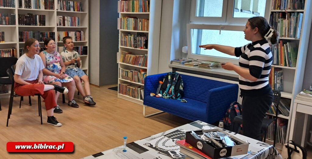 Na zdjęciu na pierwszym planie stoi dziewczyna w wieku 12-14 lat  (sweter z czarno-białe poziome grubsze pasy z rękawami kończącymi się przed łokciem) wskazująca na siedzącą naprzeciwko niej oddaloną od 2,5 m grupę 2 dziewczynek i 1 chłopczyka (wiek dzieci na oko 7-13). Wygląda jakby grały w kalambury lub w podobną zabawę. Na stole obok w otwraty pudełku widać jakieś elementy prawdopodobnie gry planszowej, w pobliży stoi klepsydra (prawdopodobnie z gry) odmierzająca czas. Pod oknem stoi chabrowa  sofa 2-osobowa. Akcja rozgrywa się w bibliotece (tu i tam poustawiane białe regały z książkami).