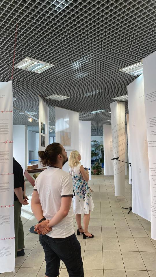 Na zdjęciu na pierwszym planie stoi zwrócony tyłem (i lekko bokiem) mężczyzna oglądający wystawę - biały t-shirt, fryzura - kok, spodnie ciemne. Ręce ma złożone z tyłu, głowa podniesiona do góry. Na dalszym planie