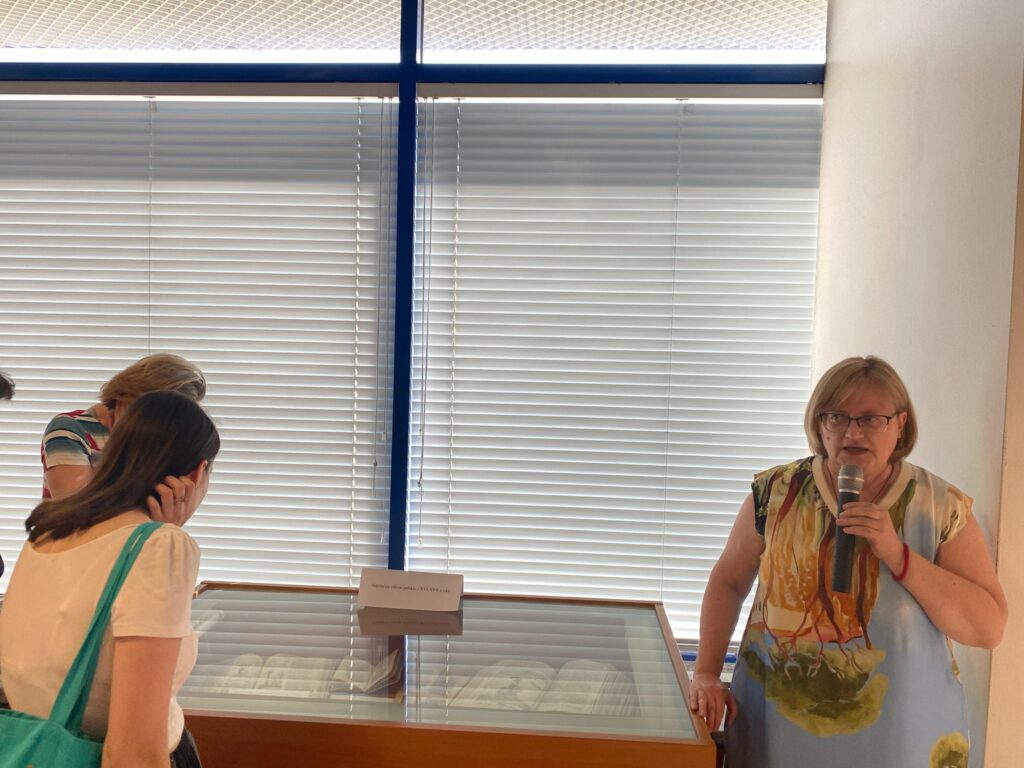 Na zdjęciu po prawej stronie kobieta mówiąca do mikrofonu, na środku gablota wystawiennicza, po lewe stojąca tyłem (a przodem do gabloty) kobieta zaglądająca do gabloty (oglądająca elementy wystawy prezentowane w poziomie w gablocie). W tle duże okno zasłonięte prze żaluzje. Kobitea trzymająca mikrofon (bezprewodowy), posiada włosy prawie do ramion, raczej jaśniejsze, założoną ma sukienkę w duże kolorowe wzory a raczej jakieś malowidło (wygląda jak budynek i drzewa drzewa na tle nieba), na nosie kobitea nosi okulary (kształt raczej prostokątny). Kobieta zaglądająca do gabloty ma białą bluzkę z krótkim rękawem, na ramieniu turkusową torbę zakupową, odgarnia włosy za ucho. Za nią widać fragment jeszcze jednej kobiety.