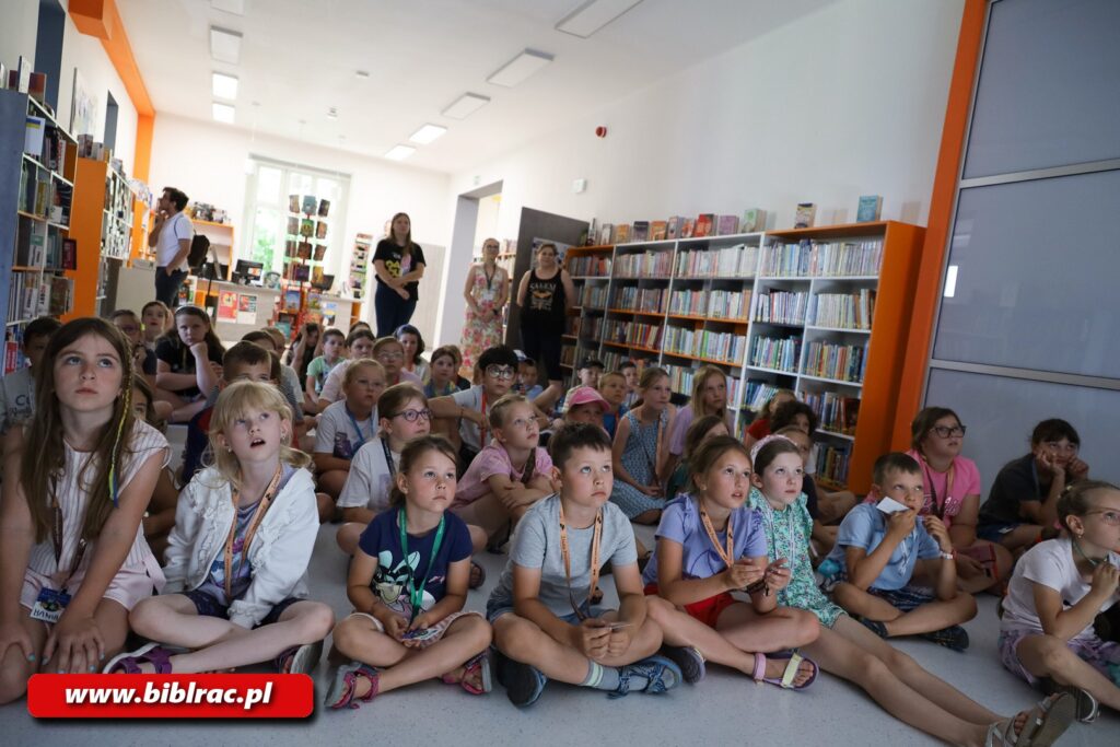 Na zdjęciu grupa dzieci w wieku siedzących na podłodze w bibliotece. Wszystkie zwrócone w jedną stronę, sprawiając wrażenie, jakby oglądały wyświetlany film lub prezentację (nie widać ekranu, gdyż znajduje się on prawdopodobnie za osobą, która robi zdjęcie). Za dziećmi w tle widać 3 kobiety - nauczycielki lub bibliotekarki. Obok kobiet stoi mężczyzna, który przegląda książki na regale. Siedzące dzieci są w strojach letnich (krótkie spodenki), mają miny, jakby były pochłonięte przez seans. Na szyjach mają tzw. smycze, prawdopodobnie z doczepionymi identyfikatorami.
