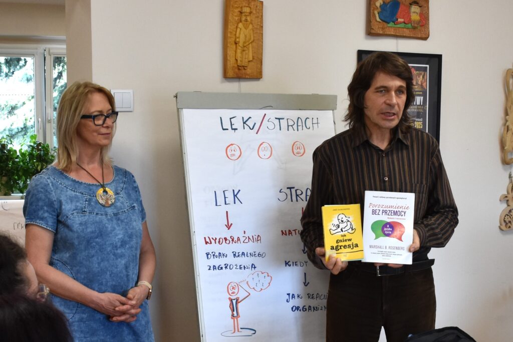 Na zdjęciu widać stojących przy tablicy (flipcharcie) kobietę i mężczyznę. Kobieta jest w średnim wieku, ma blond włosy (rozpuszczone), okulary z czarnymi oprawkami, ma na sobie niebieską sukienkę, na szyi naszyjnik z dużym okrągłym wisiorkiem z namalowanym drzewem. Ma złożone dłonie. Mężczyzna obok jest w średnim wieku, ma długie ciemne włosy (długość mniej więcej do ramion), koszulę w pionowe brązowe paski, brązowe spodnie (z czarnym paskiem). Mężczyzna trzyma w dłoniach, pokazując dwie książki, na okładkach widać tytuły "Lęk, gniew, agresja" oraz "Porozumienie bez przemocy"