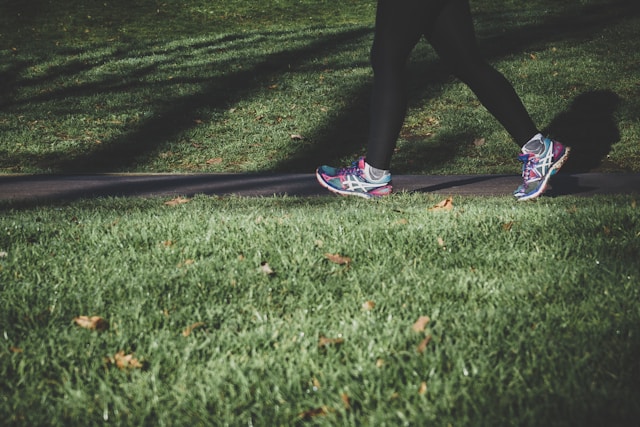 Na zdjęciu nogi osoby (chyba kobiety_od ud w dół. Legginsy sportowe i buty do biegania (różowo-niebieskie), widać, że biegnie lub szybko idzie. Bieg odbywa się po alejce (asfaltowej lub betonowej), po bokach której znajduje się trawnik. Nogi zajmują ok 1/4 kadru (w prawej górnej części).