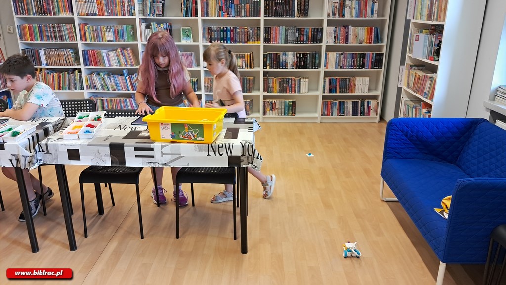 Na zdjęciu pomieszczenie biblioteki, widać białe stoły (w tym jeden tylko częściowo) a na nich porozkładane klocki Lego. Przy stołach stoją dzieci w wieku 7-9 lat ( w sumie troje - 2 dziewczynki, jeden chłopak) składające klocki. Po prawej stronie widać granatową sofę 2-3 osobową. Na dalszym planie widać regały z książkami.
