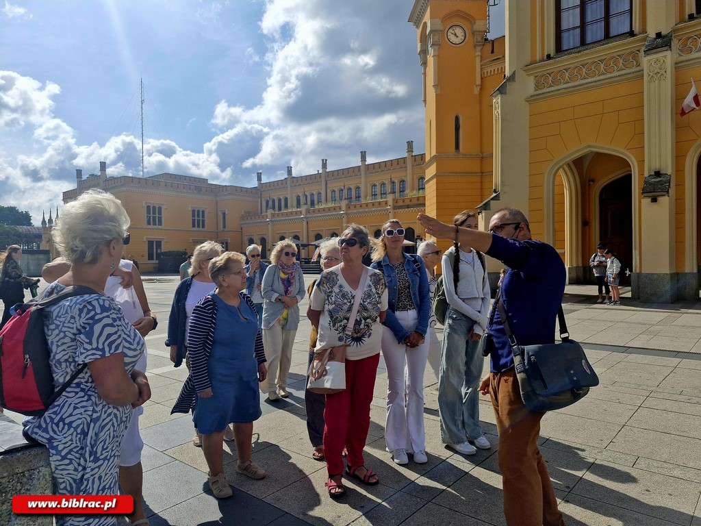 Na zdjęciu grupa pań w różnym wieku na placu przed dużym żółtym budynkiem (pałacem?). Przed paniami stoi mężczyzna wyglądający na przewodnika. Wskazuje palcem w lewo. Nosi granatową koszulę 