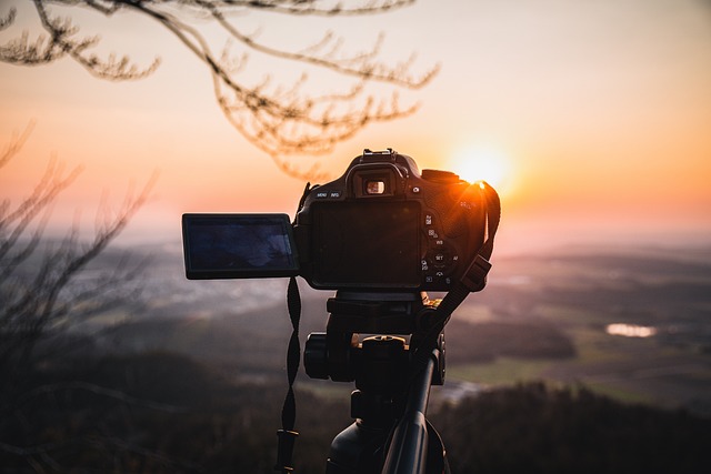 Na zdjęciu aparat fotograficzny na statywie, sfotografowany od tyłu. W dali, przed aparatem, krajobraz (pagórki, drzewa, zachodzące słońce).