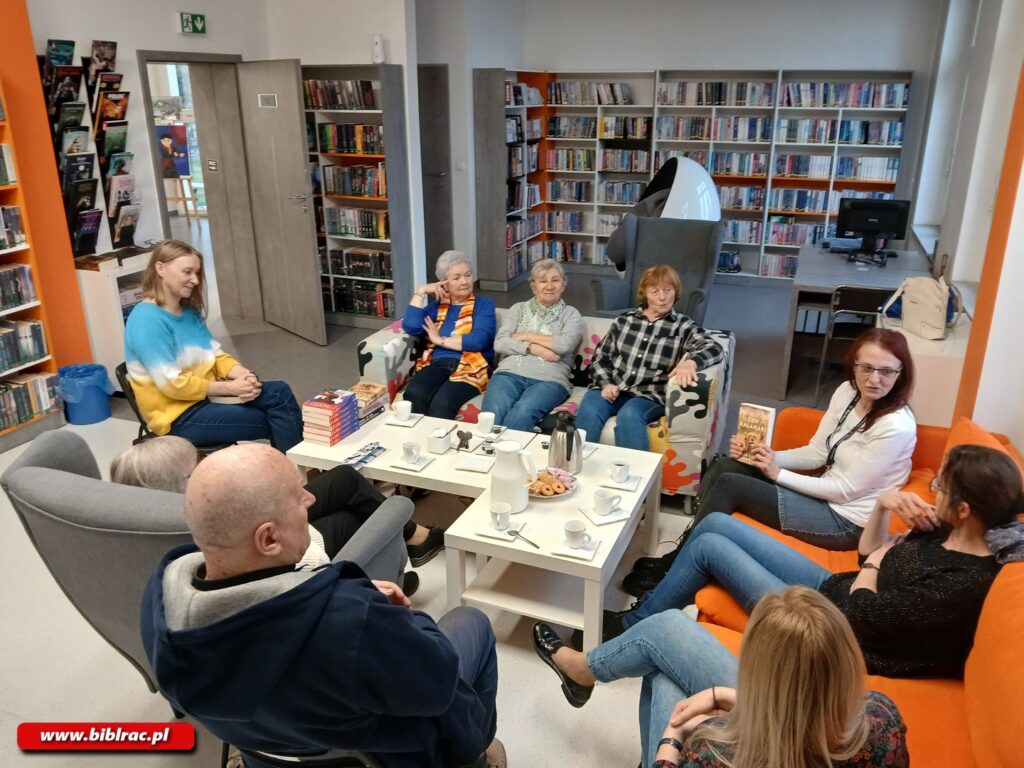 Na zdjęciu osiem dorosłych osób w różnym wieku siedzących na fotelach i kanapach w bibliotece wokół stolika kawowego.