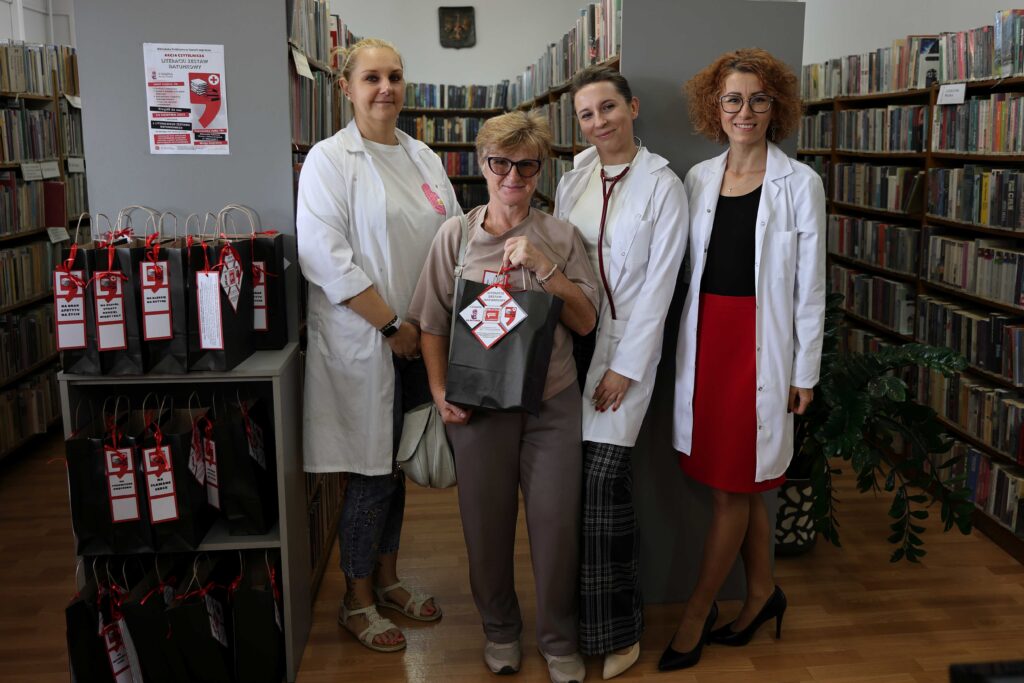 Na zdjęciu pośród regałów grupa 4 bibliotekarek przebranych za medyków. Jedna trzyma w ręce zapakowany zestaw książek