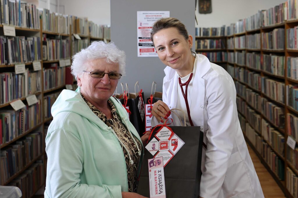 Na zdjęciu pośród regałów bibliotecznych stoją dwie kobiety - bibliotekarka przebrana za medyka oraz czytelniczka (starsza pani). Wspólnie trzymają w ręce pakunek.