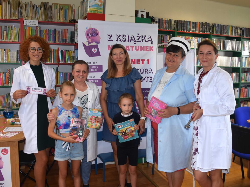 Na zdjęciu bibliotekarki i czytelnicy (kobieta i dzieci - chłopczyk i dziewczynka). Bibliotekarki są przebrane za medyków. Wszyscy pozują do zdjęcia. Dzieci trzymają prezenty - książeczki.