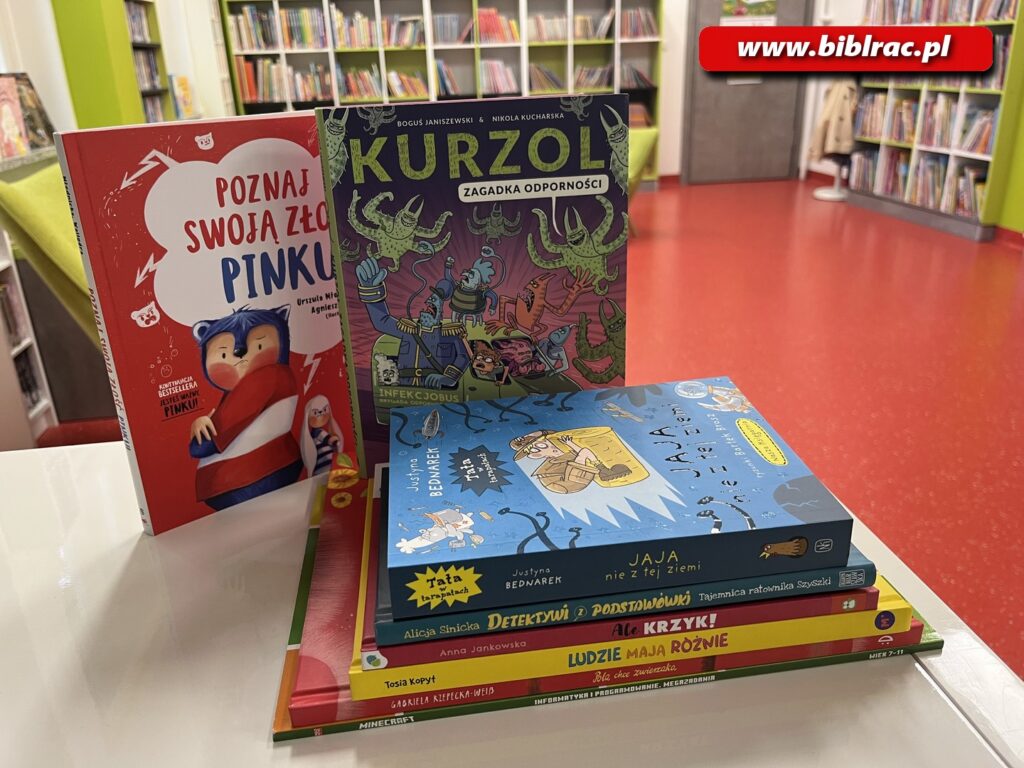 Na zdjęciu 6 książek ułożonych w stosie na jasnym stole oraz dwie książki stojąca za tym stosem. W tle widać czerwoną podłogę oraz biało-szaro-limonkowe regały biblioteczne.