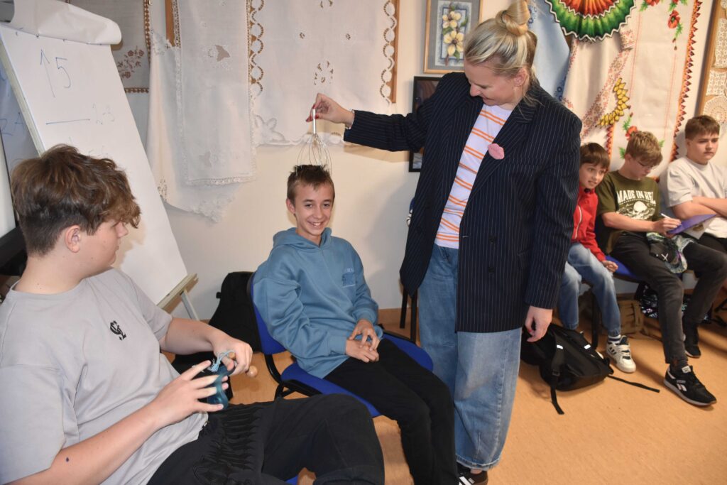 Na zdjęciu chłopcy siedzący na krzesłach oraz pani prowadząca (w luźnej marynarce oraz bluzce w czerwone paski) masująca po głowie jednego z chłopców urządzeniem drucikowym do masażu (chłopak się śmieje).