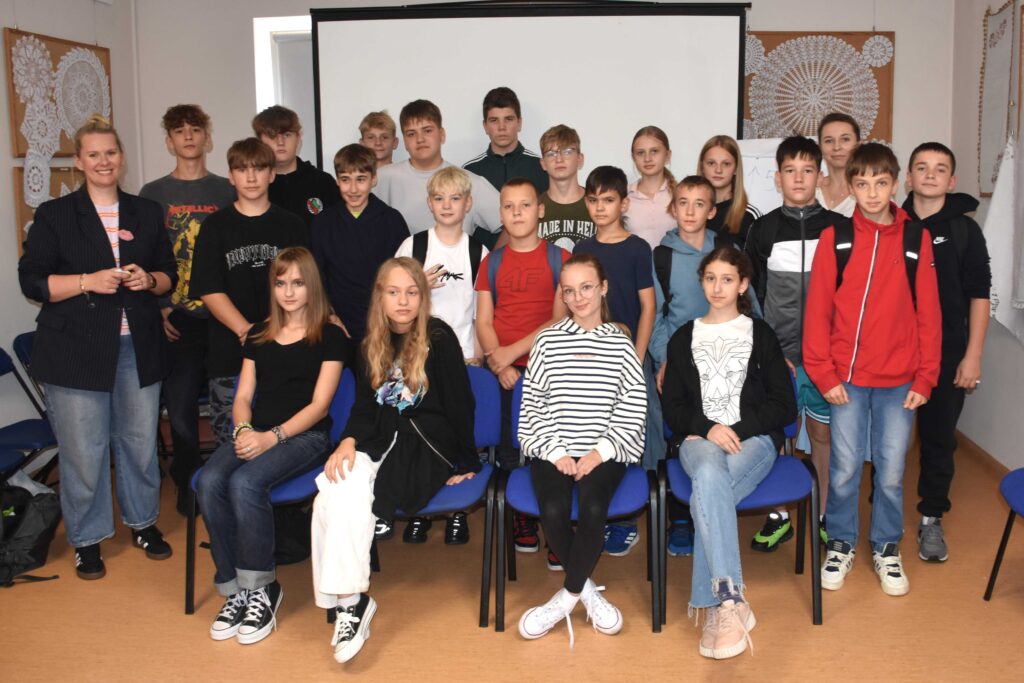 Zdjęcie grupowe zrobione prawdopodobnie w bibliotece przedstawiające młodzież (i dzieci) oraz panią prowadzącą warsztaty (na zdjęciu pozuje z lewej strony)