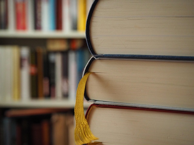 Na zdjęciu mocne zbliżenie na stos 3 książek (widać fragmenty tych książek). Z jednej książki wystaje wstążka. W oddali, w tle widać rozmazane regały z książkami