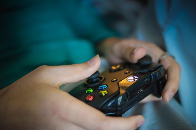 Na zdjęciu zbliżenie na dłonie młodej osoby trzymającej gamepad konsoli Xbox. Urządzenie ma kolor czarny, przyciski są w kolorach: czerwonym, zielonym, niebieskim i żółtym. Na gamepadzie znajduje się podświetlane logo (zniekształcona litera "X" w kółku).