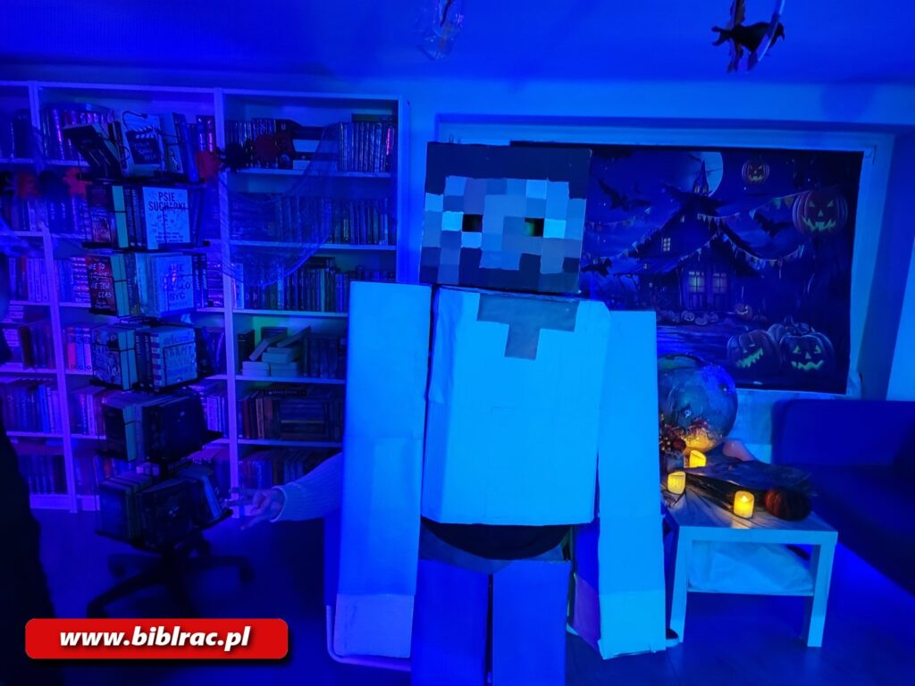 Na zdjęciu pomieszczenie biblioteczne w świetle fioletowym (półmrok), widać na pierwszym planie kogoś (prawdopodobnie dziecko) przebranego za postać z gry komputerowej Minecraft