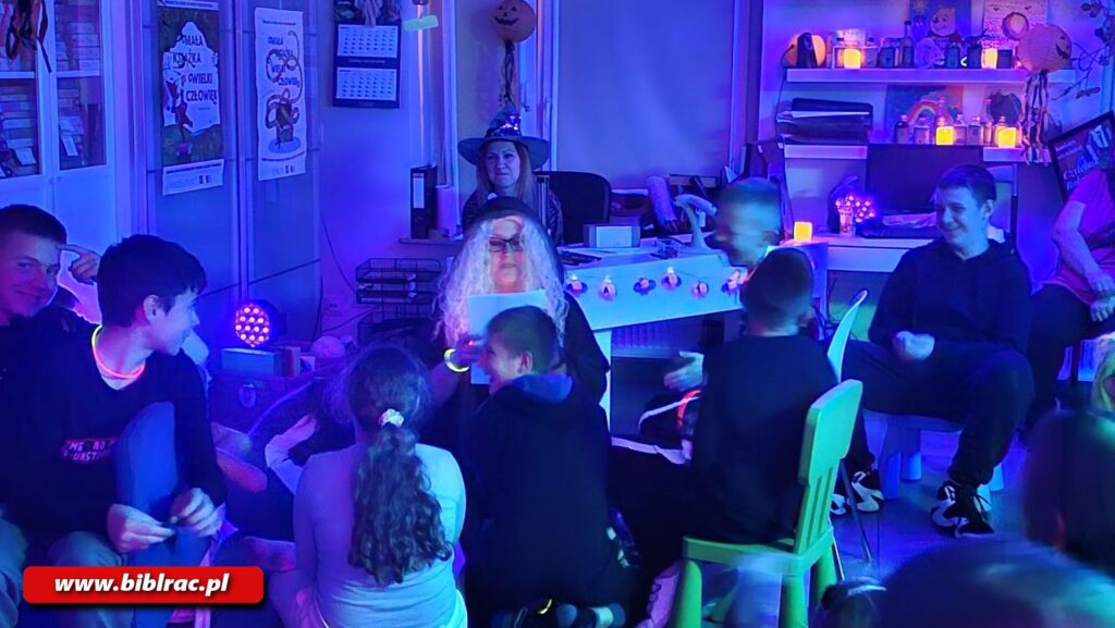 Na zdjęciu pomieszczenie biblioteczne w świetle fioletowym (półmrok), widać dzieci podczas imprezy halloweenowej