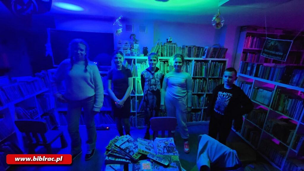Na zdjęciu pomieszczenie biblioteczne w świetle fioletowym (półmrok), widać dzieci podczas imprezy halloweenowej