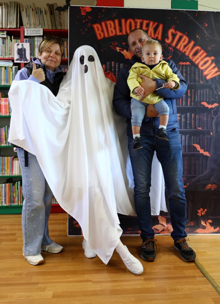 Grupa trzech dorosłych osób i małe dziecko pozuje do zdjęcia na tle planszy z napisem „Biblioteka Strachów”. Jedna osoba jest przebrana za ducha w białym prześcieradle z czarnymi oczami i ustami, dwie inne uśmiechają się i stoją obok, a mężczyzna trzyma na rękach małego chłopca w żółtej kurtce. W tle widać półki z książkami i dekoracje w halloweenowym stylu.