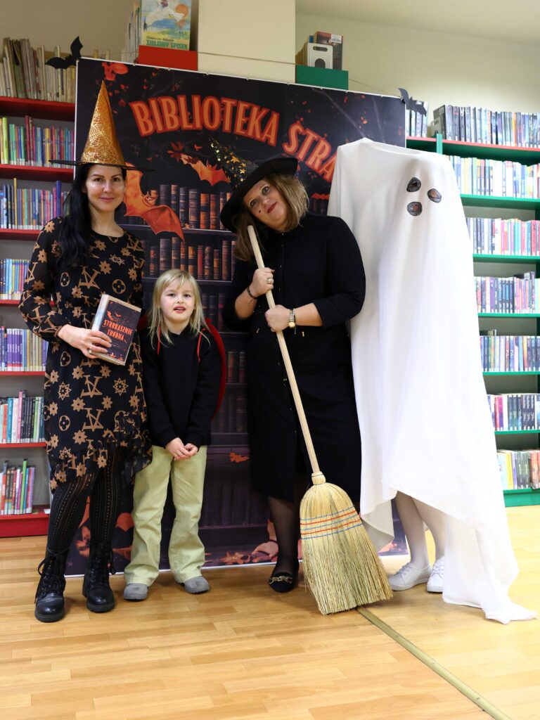 Dwie kobiety i dziecko pozują z osobą w przebraniu ducha na tle planszy „Biblioteka Strachów”. Dorosłe kobiety mają na głowach czarodziejskie kapelusze, jedna trzyma miotłę. Dziewczynka uśmiecha się, stojąc między nimi. W tle widać regały z książkami i jesienno-halloweenowe ozdoby.