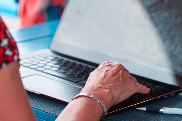 Na zdjęciu widać ramię i dłoń starszej osoby spoczywająca na klawiaturze otwartego laptopa. Na nadgarstku widać srebrną bransoletkę.