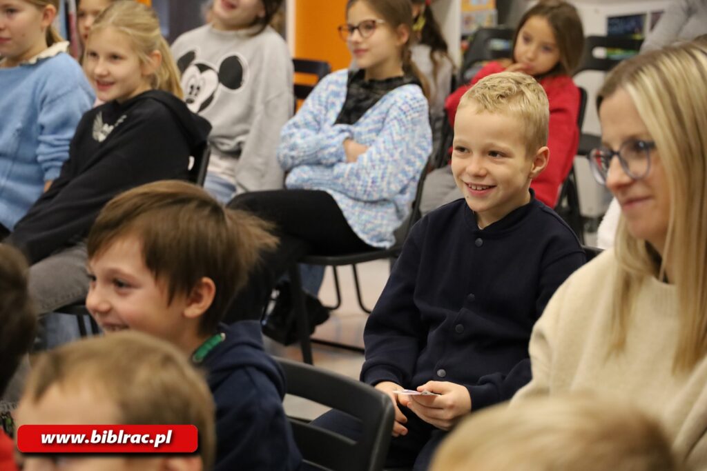 Zbliżenie na dzieci siedzące na widowni. Wszystkie dzieci uśmiechnięte. Po prawej stronie kadru młoda kobieta w okularach, również uśmiechnięta,