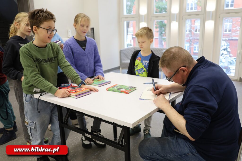 Dzieci stojące przy białym stole czekające na autograf, mężczyzna w okularach, z krótkimi włosami podpisujący książki,.