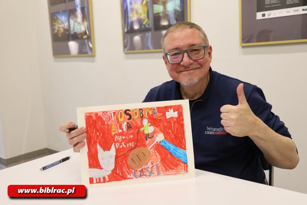 Na zdjęciu pozuje uśmiechnięty mężczyzna siedzący przy białym stole. Jedną ręką trzyma ilustrację lub książkę i pokazuje ją do aparatu, Drugą ręką pokazuje podniesiony do góry kciuk.