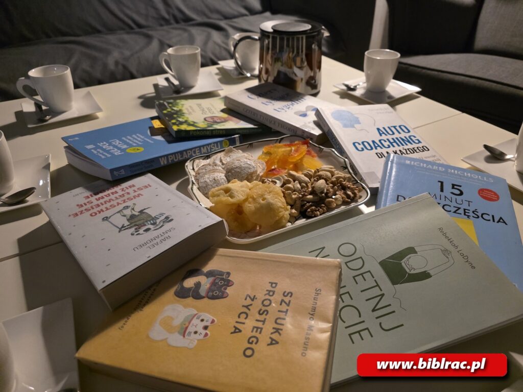 Na zdjęciu stół, na którym porozkładane są różne książki, przekąski na kwadratowym talerzu, kubki do picia herbaty. 