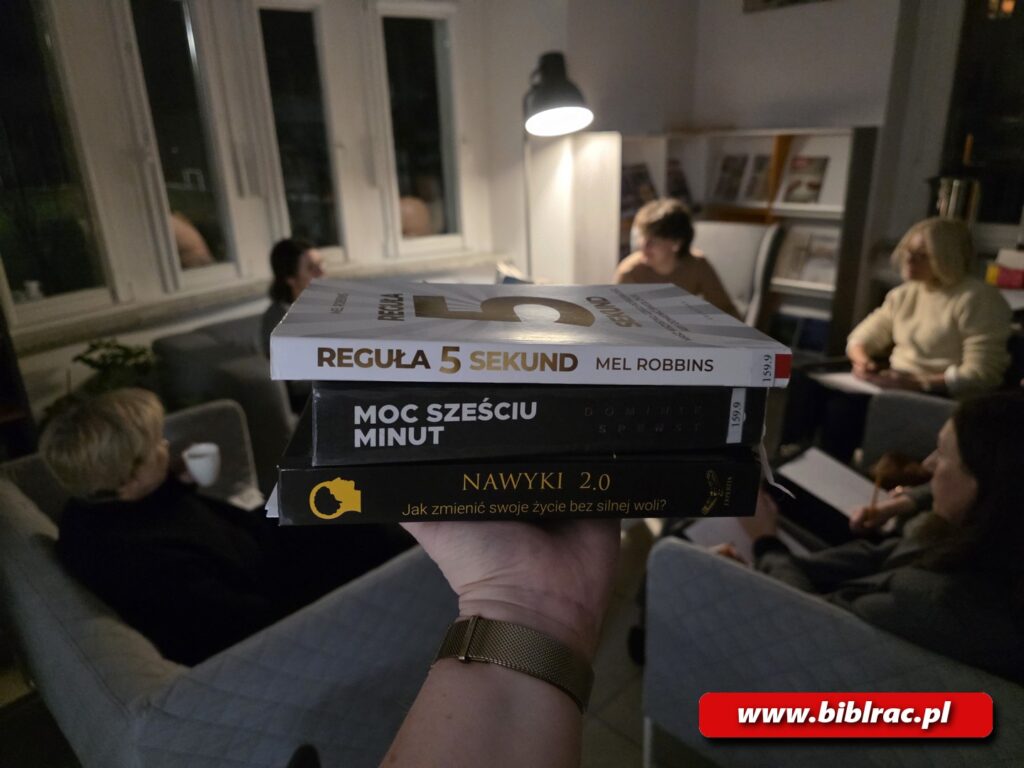 Na zdjęciu dłoń trzymająca stos książek, w tle osoby na siedzące fotelach przy świetle lampy stojącej, prawdopodobnie  w bibliotece