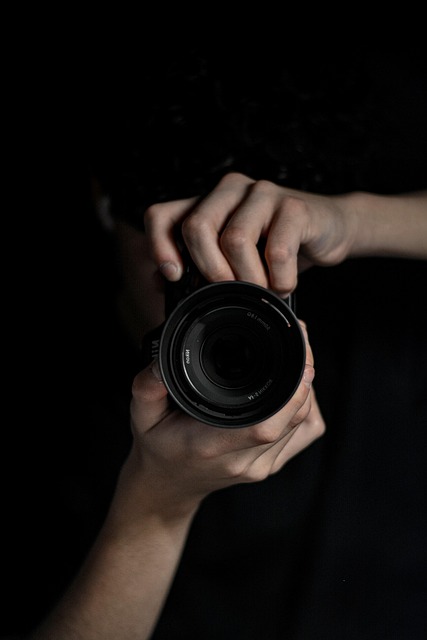 Zdjęcie wykonane w pionie, przedstawiające aparat fotograficzny (na środku kadru), skierowany obiektywem na wprost (czyli w stronę odbiorcy), trzymany przez mężczyznę (jego twarz schowana za aparatem), tło na zdjęciu jest czarne, koszulka osoby trzymającej aparat również czarna.