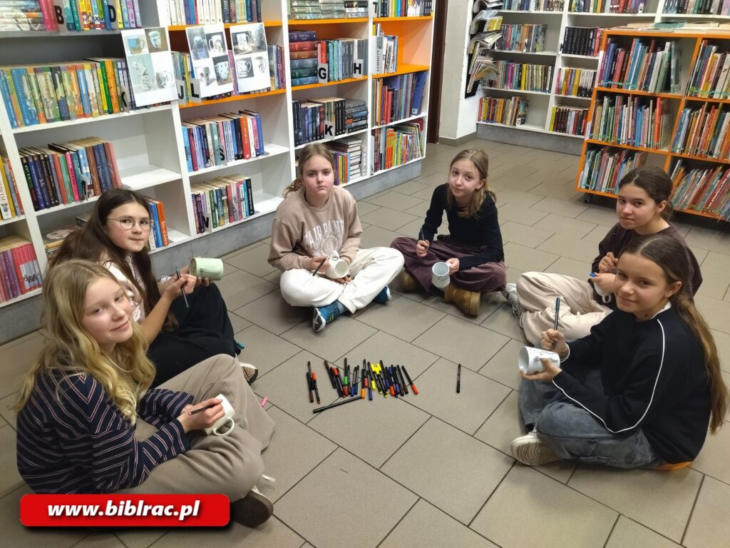 Na zdjęciu grupa 6 dziewczyn w wieku 12-15 lat siedzących w półokręgu po turecku na kafelkach. Na środku podłogi leży kilkadziesiąt pisaków. Dziewczyny trzymają w rękach kubki z namalowanymi obrazkami i spoglądają w obiektyw.