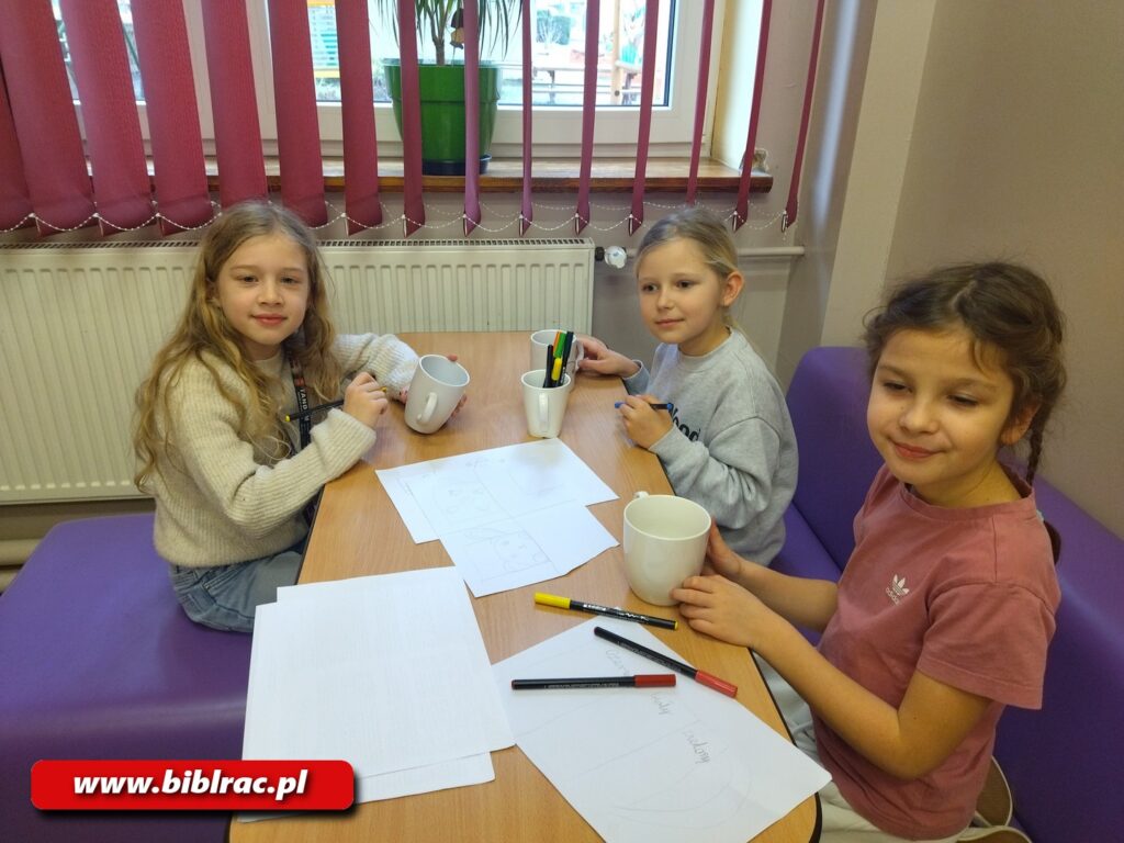 Na zdjęciu podłużny stół (wyglądający jak szkolna ławka), a przy nim trzy dziewczynki w wieku 7-9 lat. Każda z nich spogląda w obiektyw i uśmiechają się, zdjęcie zostało wykonane w czasie procesu twórczego - dziewczyny malują wzory na ceramicznych kubkach.