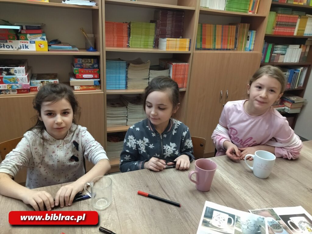 Na zdjęciu podłużny stół , a przy nim trzy dziewczynki w wieku 7-10 lat. Każda z nich spogląda w obiektyw i uśmiechają się, zdjęcie zostało wykonane w czasie procesu twórczego - dziewczyny malują wzory na ceramicznych kubkach.