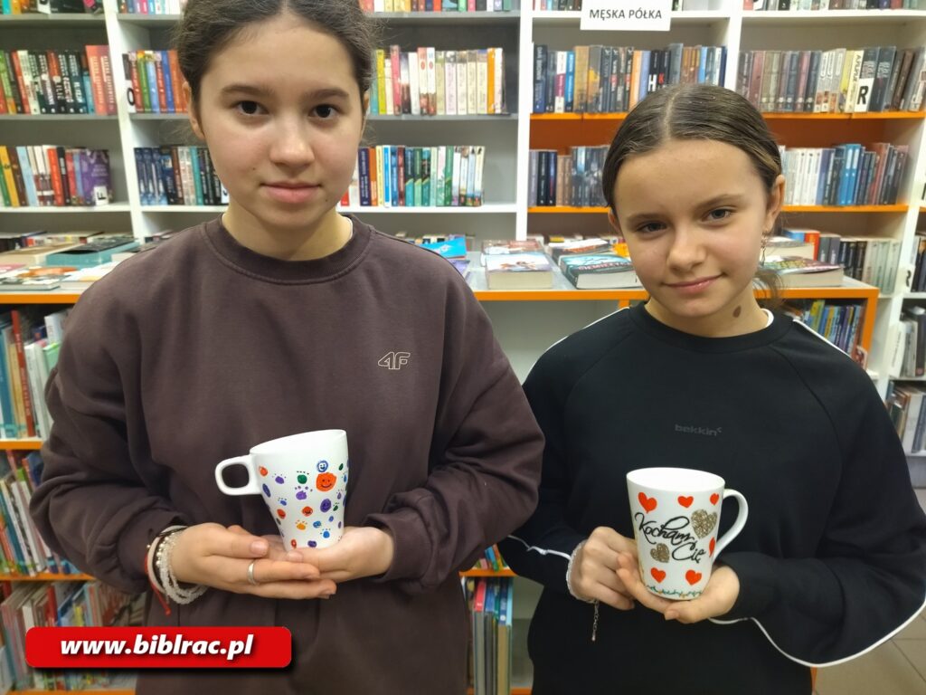 Dwie dziewczyny w wieku 8-12 lat trzymające kubki ceramiczne z namalowanymi wzorami. W tle regały z książkami.