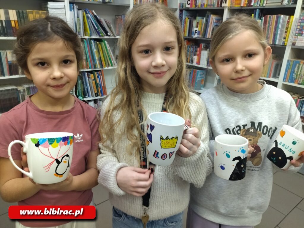 Na zdjęciu pozujące 3 dziewczynki w wieku 7-9 lat, każda trzymająca w ręku biały kubek z namalowanym wzorem (dziewczynka po prawe trzyma 2 kubki). Dziewczynka po lewej (szatynka z upiętymi włosami) ma na kubku namalowanego pingwina z balonikami, dziewczynka w środku (blondynka z rozpuszczonymi włosami) żółtą koronę i napis Love, dziewczynka po prawej (blondynka) trzyma kubki z namalowanymi czarnymi zwierzątkami (prawdopodobnie pieskami). W tle regały z książkami. 