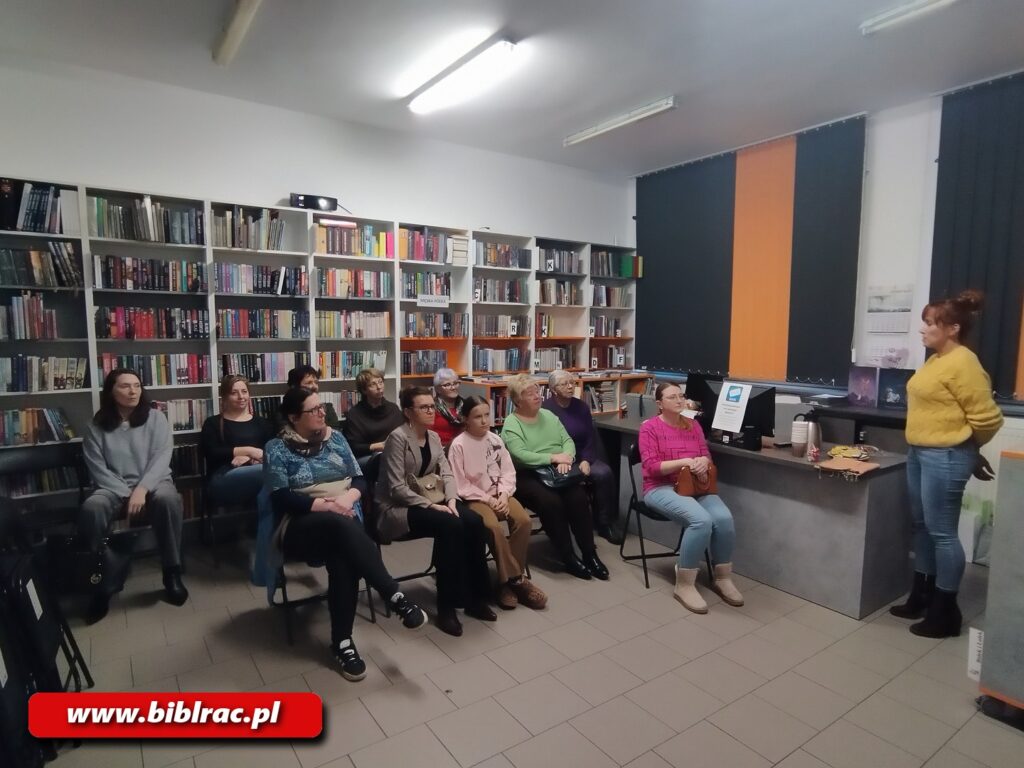 Na zdjęciu kobieta w żółtym swetrze i dżinsach przemawia do grupy ludzi siedzących na krzesłach w pomieszczeniu bibliotecznym. W tle rgały z książkami