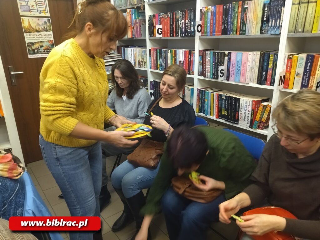 Na zdjęciu kobieta w żółtym swetrze i dżinsach rozdająca kobietom siedzącym na krzesłach jakieś saszetki.