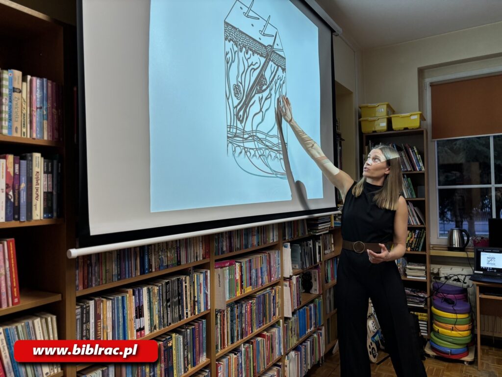 Na zdjęciu młoda kobieta ubrana na czarno (bluzka, spodnie lub jest to kombinezon, do tego brązowy szeroki pas, pokazuje coś na ekranie, na który obraz rzuca projektor. 