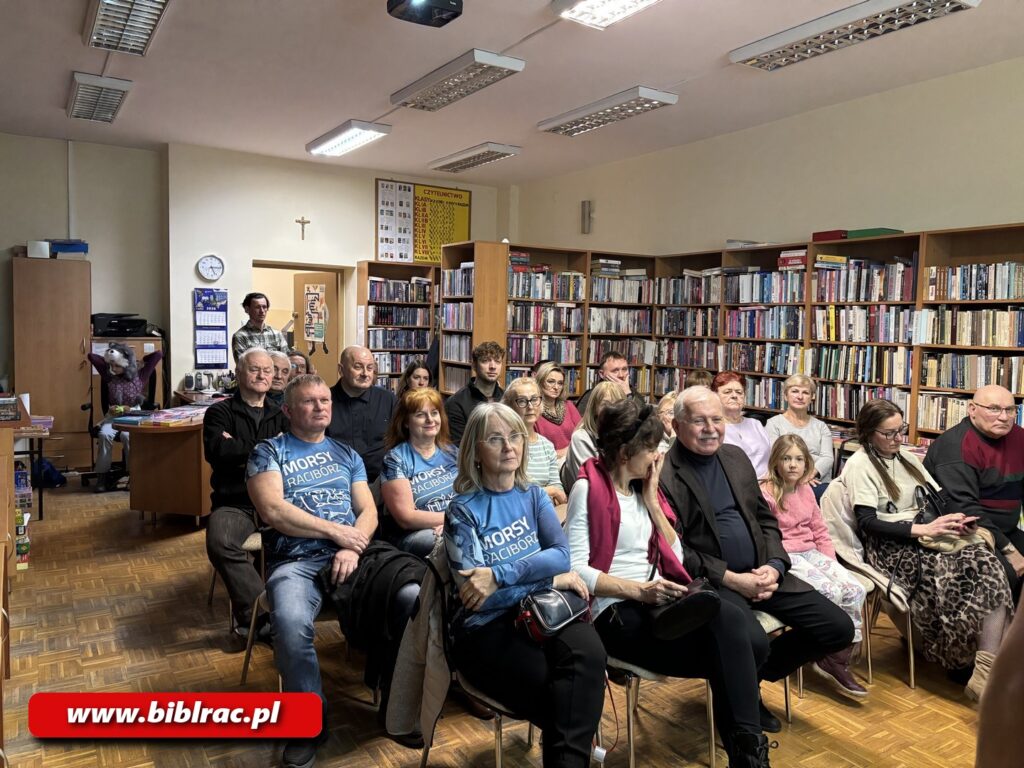 Na zdjęciu pomieszczeni biblioteczne, widać regały z książkami oraz kilkanaście krzeseł ustawionych dla widowni. Krzesła zapełnione są przez ludzi w różnym wieku spoglądających przed siebie.