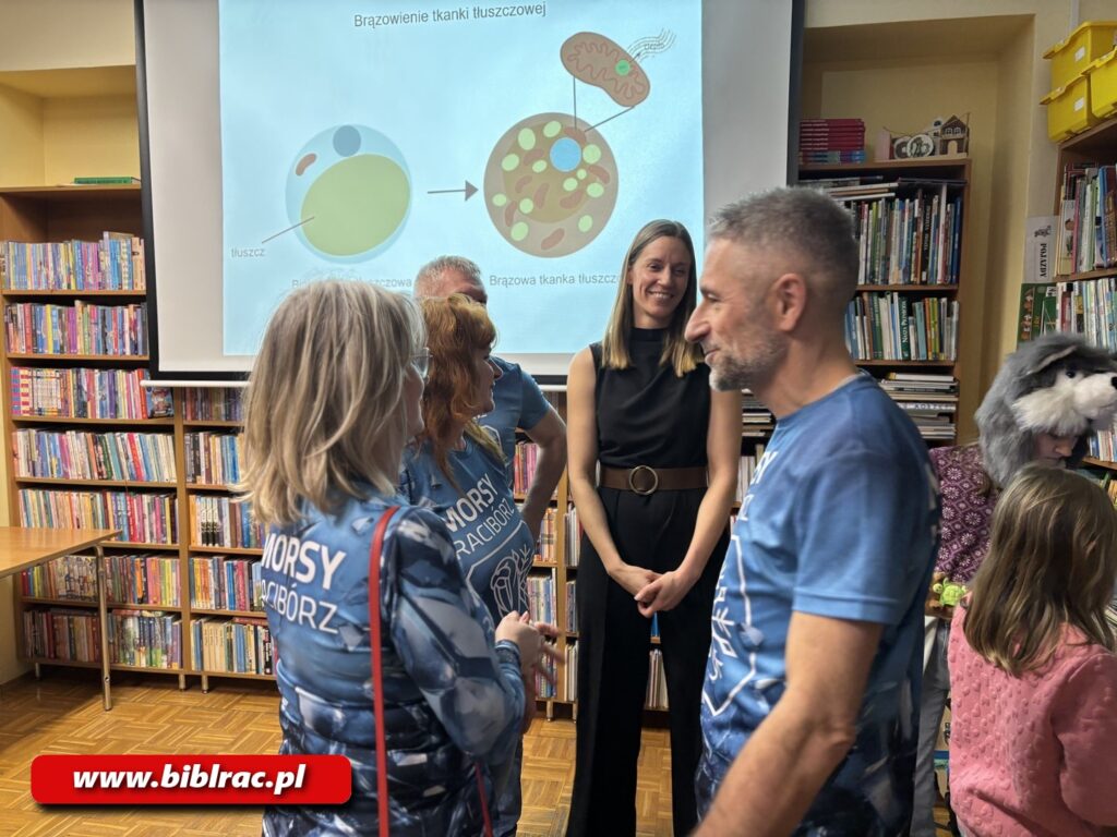 Na zdjęciu pomieszczenie biblioteczne, w tle rozwinięty ekran z rzucnonym n aniego wobrazem z projektowra przedstawiającym jakies okrągłe okręgi i legendę do nich. Poza tym widać przechodzącego mężczyznę w średnim wieku z krótką siwą brodą, w niebieskim t-shircie, obok niego stoi kobieta odwrócona tyłem do aparatu, gdzieś dalej stoi uśmiechnięta młoda kobieta (blondynka z włosami nieco poza ramiona) ubrana na czarno (kombinezon, do tego brązowy szeroki pas).