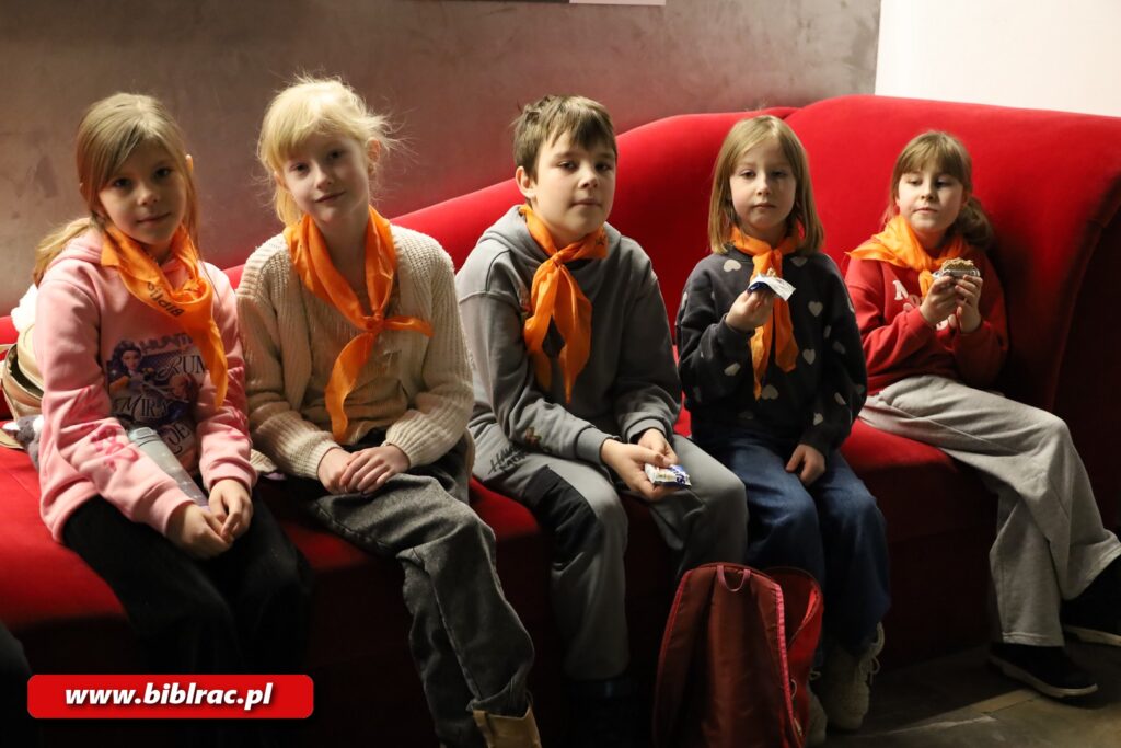 Na zdjęciu 5 dzieci siedzących na czerwonym siedzisku i pozujących do zdjęcia. Na szyjach mają założone pomarańczowe chusty.