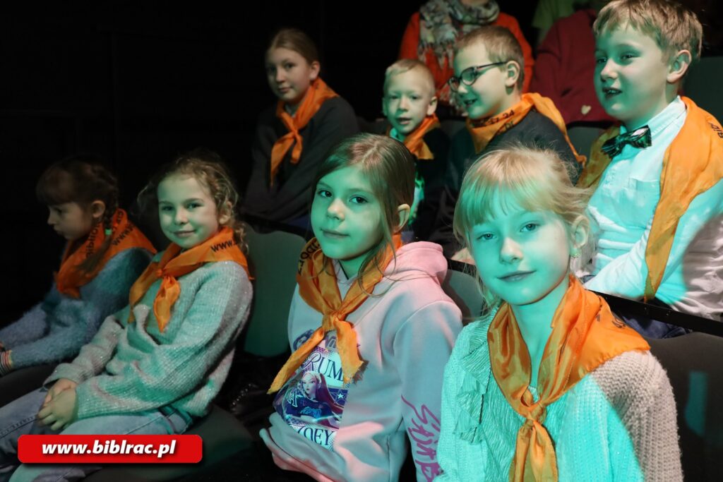 Na zdjęciach zbliżenie na dzieci siedzące na widowni, mają założone na szyjach pomarańczowe chusty,, spoglądają w stronę aparatu, pada na nie niebieskozielone światło.